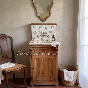 May include: A wooden vanity cabinet with a floral patterned backsplash and sink basin, featuring a gold-framed mirror above it. To the left is a wooden chair with a woven seat, and to the right is a wicker basket. The text Collectiondemaman is visible.