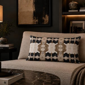 May include: A rectangular throw pillow featuring a symmetrical, repeating geometric pattern in shades of black, cream, and tan. The pillow is positioned on a light beige sofa, showcasing a distinct, bold, and intricate design across its surface.