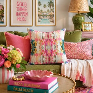 May include: A square throw cushion featuring a symmetrical abstract pattern in shades of pink, green, white, and gold. The cushion is displayed on a green sofa alongside a solid pink cushion and a rectangular pink patterned cushion with teal trim.