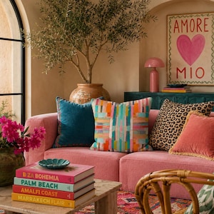May include: A pink sofa featuring four decorative throw pillows in teal, multicolored stripes, leopard print, and solid coral with fringe. A stack of four books titled B ZA BOHEMIA, PALM BEACH, AMALFI COAST, and MARRAKECH FLAIR sits on a stone table.