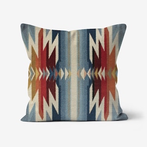 May include: Square throw pillow with a geometric pattern in shades of blue, red, cream, and gold. The design features vertical stripes and zigzag shapes, creating a Southwestern or Native American inspired aesthetic. The pillow is likely made of a woven fabric.