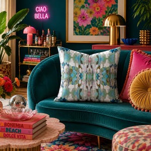 May include: A square throw pillow featuring a symmetrical abstract pattern in shades of teal, blue, green, and white. The pillow is positioned on a dark teal velvet sofa alongside a solid pink pillow and a round, pleated mustard yellow pillow.