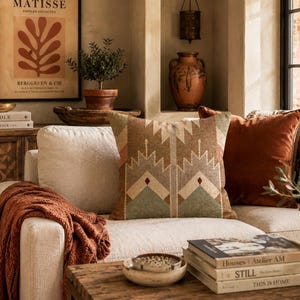 May include: A square throw pillow featuring a geometric pattern in shades of brown, tan, sage green, and cream. The pillow is placed on a light-colored sofa alongside a solid rust-colored velvet pillow and a textured rust-colored throw blanket.