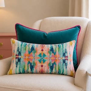 May include: A rectangular lumbar pillow featuring a multicolored abstract brushstroke pattern in shades of teal, pink, orange, and yellow, layered in front of a solid teal velvet pillow with a bright pink piped edge.