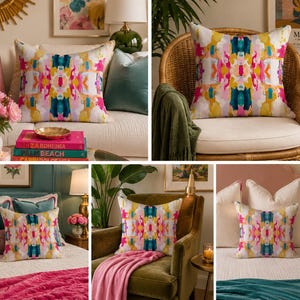 May include: A square throw pillow featuring a vibrant, symmetrical abstract pattern with bold splashes of magenta, teal, mustard yellow, and white. The design displays a mirrored, painterly aesthetic across the fabric surface.
