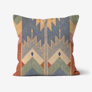 May include: Square throw pillow with a geometric pattern in shades of blue, yellow, green, and rust. The design features stylized arrow and diamond shapes, creating a textured appearance. The pillow has a soft, inviting look.