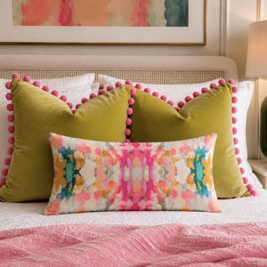 May include: Two square olive green velvet throw pillows with pink pom-pom trim and one rectangular lumbar pillow featuring a multicoloured abstract watercolour print in shades of pink, teal, orange, and white, arranged on a white bedspread.