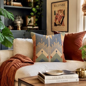 May include: A square throw pillow featuring a geometric pattern in shades of blue, cream, and rust orange. The pillow is placed on a white sofa next to a solid rust-colored velvet pillow, a textured orange throw blanket, and a stack of books.