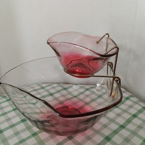 Op de afbeelding: Een glazen serveerschaal met twee verdiepingen en een metalen standaard. De bovenste kom is roze en de onderste kom is helder met een roze rand.