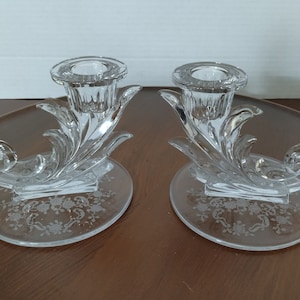 May include: A pair of clear glass candle holders. Each holder features a circular base with an etched floral design, supporting a decorative, swirling stem that holds a candle cup. The design is elegant and classic.