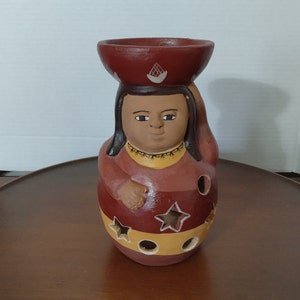 Peut inclure: Un bougeoir en céramique en forme de femme portant un bol sur la tête. La femme est peinte dans des tons de brun, rouge et jaune avec un motif d'étoiles.