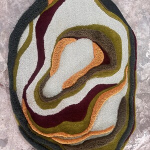 This image features a unique, irregularly shaped tufted area rug lying flat on a light oak wood floor in a cozy, modern bedroom. The rug has an organic, freeform silhouette with wavy edges, resembling a topographical map or an abstract geode slice. It boasts a deeply textured, high-low pile with swirling, 3D carved bands of color that create a tactile, contoured look