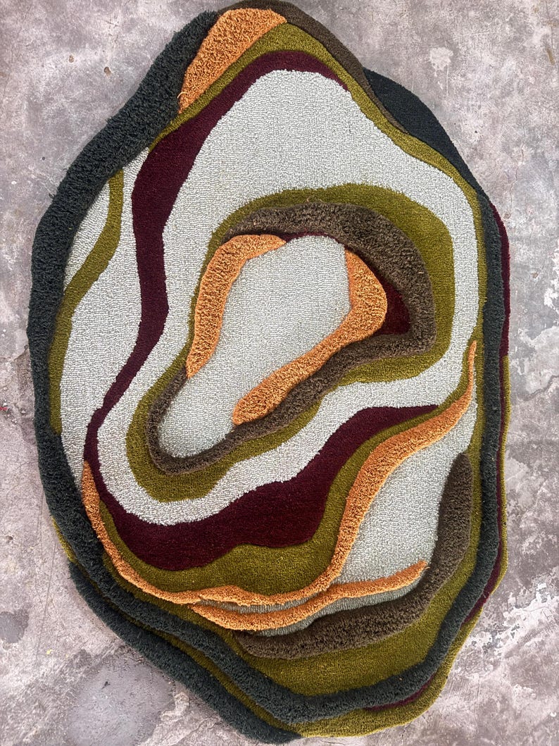 This image features a unique, irregularly shaped tufted area rug lying flat on a light oak wood floor in a cozy, modern bedroom. The rug has an organic, freeform silhouette with wavy edges, resembling a topographical map or an abstract geode slice. It boasts a deeply textured, high-low pile with swirling, 3D carved bands of color that create a tactile, contoured look