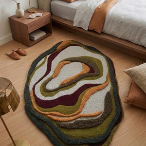 This image features a unique, irregularly shaped tufted area rug lying flat on a light oak wood floor in a cozy, modern bedroom. The rug has an organic, freeform silhouette with wavy edges, resembling a topographical map or an abstract geode slice. It boasts a deeply textured, high-low pile with swirling, 3D carved bands of color that create a tactile, contoured look