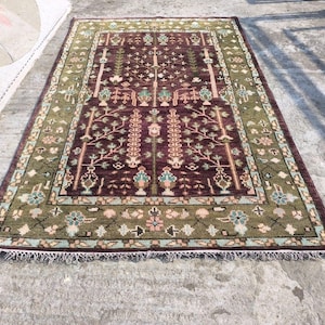 May include: A rectangular area rug with a central burgundy field and a green border. The rug features a detailed floral and tree design in shades of beige, blue, and pink. The rug has a fringe along the edges.