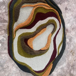 This image features a unique, irregularly shaped tufted area rug lying flat on a light oak wood floor in a cozy, modern bedroom. The rug has an organic, freeform silhouette with wavy edges, resembling a topographical map or an abstract geode slice. It boasts a deeply textured, high-low pile with swirling, 3D carved bands of color that create a tactile, contoured look