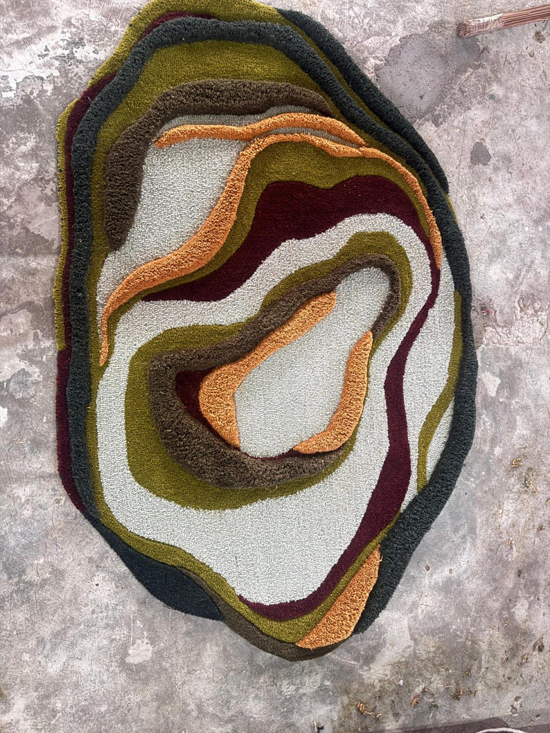 This image features a unique, irregularly shaped tufted area rug lying flat on a light oak wood floor in a cozy, modern bedroom. The rug has an organic, freeform silhouette with wavy edges, resembling a topographical map or an abstract geode slice. It boasts a deeply textured, high-low pile with swirling, 3D carved bands of color that create a tactile, contoured look