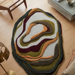 This image features a unique, irregularly shaped tufted area rug lying flat on a light oak wood floor in a cozy, modern bedroom. The rug has an organic, freeform silhouette with wavy edges, resembling a topographical map or an abstract geode slice. It boasts a deeply textured, high-low pile with swirling, 3D carved bands of color that create a tactile, contoured look