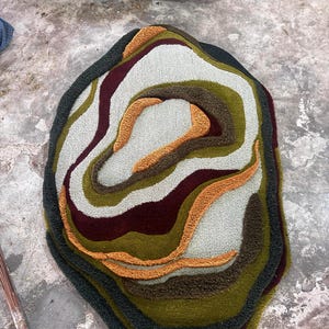 This image features a unique, irregularly shaped tufted area rug lying flat on a light oak wood floor in a cozy, modern bedroom. The rug has an organic, freeform silhouette with wavy edges, resembling a topographical map or an abstract geode slice. It boasts a deeply textured, high-low pile with swirling, 3D carved bands of color that create a tactile, contoured look