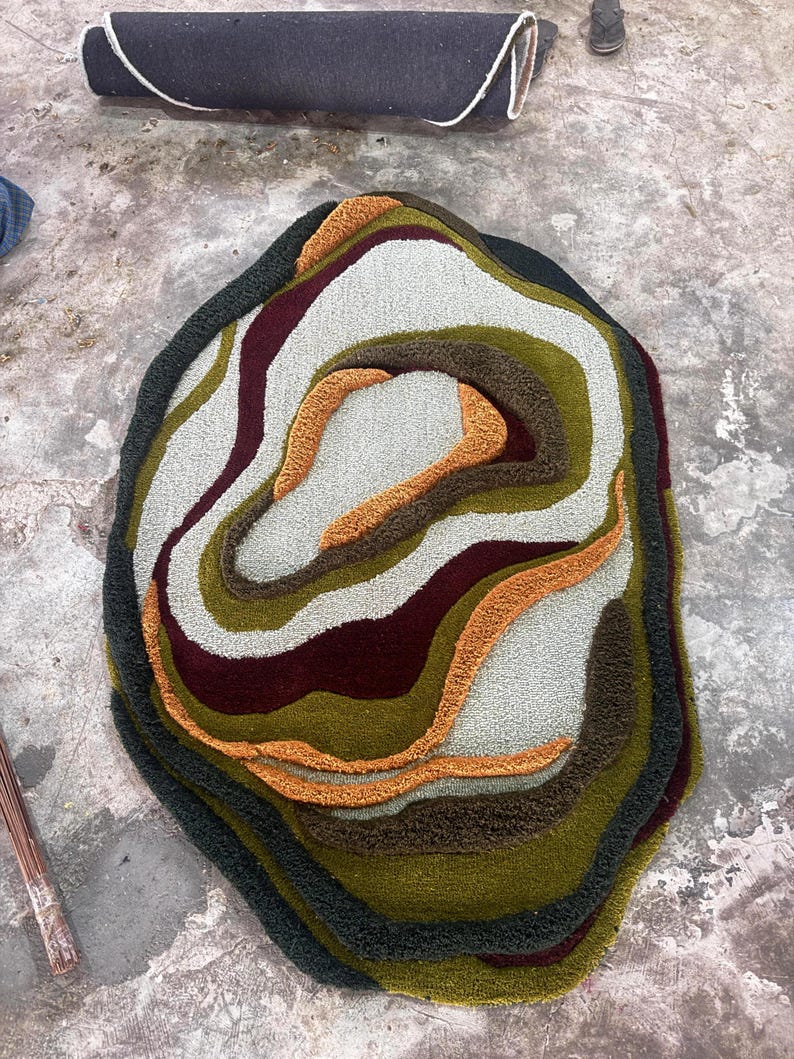 This image features a unique, irregularly shaped tufted area rug lying flat on a light oak wood floor in a cozy, modern bedroom. The rug has an organic, freeform silhouette with wavy edges, resembling a topographical map or an abstract geode slice. It boasts a deeply textured, high-low pile with swirling, 3D carved bands of color that create a tactile, contoured look