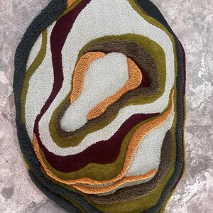 This image features a unique, irregularly shaped tufted area rug lying flat on a light oak wood floor in a cozy, modern bedroom. The rug has an organic, freeform silhouette with wavy edges, resembling a topographical map or an abstract geode slice. It boasts a deeply textured, high-low pile with swirling, 3D carved bands of color that create a tactile, contoured look