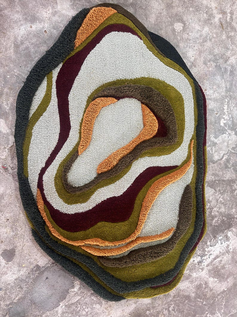 This image features a unique, irregularly shaped tufted area rug lying flat on a light oak wood floor in a cozy, modern bedroom. The rug has an organic, freeform silhouette with wavy edges, resembling a topographical map or an abstract geode slice. It boasts a deeply textured, high-low pile with swirling, 3D carved bands of color that create a tactile, contoured look