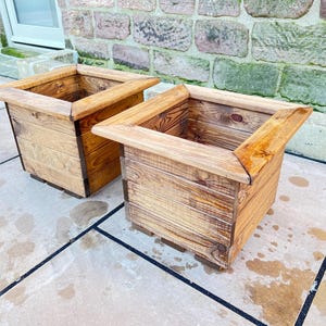 May include: Two wooden planter boxes, one in front of the other, both with a natural wood finish. The boxes are square and have a slightly raised lip around the top.