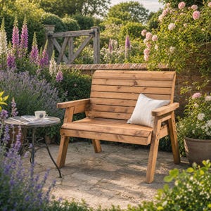 May include: A wooden garden bench with a slatted back and seat, featuring a light brown finish and sturdy armrests. A single white square throw pillow rests on the seat. Beside the bench sits a small round metal table with a cup and magazine.