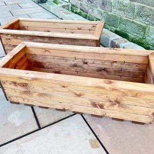 May include: Two rectangular wooden planters with a natural finish. The planters are made of wooden planks and have a slatted base. The planters are empty and ready for planting flowers or other plants. The planters are sitting on a stone surface.