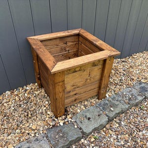 May include: A square wooden planter box with a natural brown stain finish. The planter is made of wood planks and has a simple, rustic design.