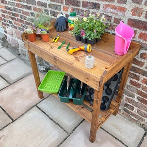 May include: A wooden potting bench with a shelf, a pink watering can, gardening tools, and plants in pots.