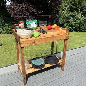 May include: A wooden serving trolley with two shelves. The top shelf has a wooden chopping board, a bowl of salad, and a bottle of wine. The bottom shelf has glasses and plates.