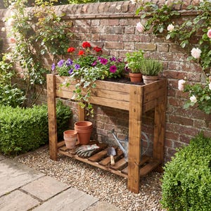 May include: A rectangular wooden raised garden bed on four legs with a lower slatted shelf. The top container holds red and purple flowers and green foliage. The lower shelf contains three terracotta pots, gardening gloves, and a metal watering can.