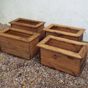 May include: Four rectangular wooden planters in a natural wood stain. Each planter features a raised border and is made from horizontal wooden planks. The planters are arranged on a bed of small pebbles, with a white wall in the background.
