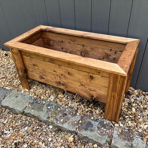 May include: A rectangular wooden planter box with a natural brown stain. The planter is made of rustic wood planks and has a raised lip around the top.