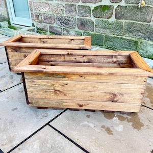 May include: Two rectangular wooden planters with a natural finish. The planters are constructed from horizontal wooden planks and feature a decorative top trim. The planters are designed for outdoor use and are placed on a stone patio.