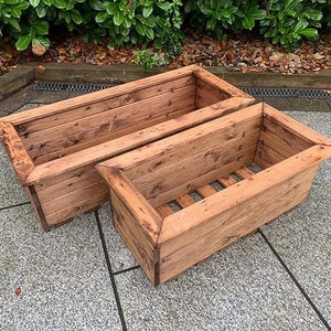 May include: Two rectangular wooden planters with a natural finish. The planters are made from horizontal wooden planks and have a slatted base for drainage. The edges are angled, and the wood grain is visible.