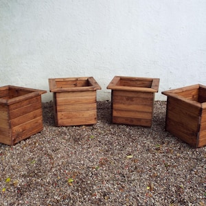 May include: Four wooden planter boxes, each with a square shape and a dark brown stain. The boxes are arranged in a row on a gravel surface.