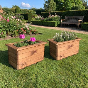 May include: Two rectangular wooden planters with a natural brown finish. The left planter contains bright pink flowers with green foliage, while the right planter holds white flowering plants. Both planters feature horizontal wood planks.