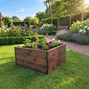 May include: A square, dark brown stained wooden planter box filled with dark soil and a variety of colourful blooming flowers, including pink, purple, and white petals, positioned on a green lawn in an outdoor garden setting.