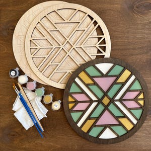 Pode incluir: Um kit de artesanato DIY com dois painéis circulares de madeira inacabados com recortes geométricos, potes de tinta, pincéis e um painel concluído. O painel concluído tem um design geométrico com seções rosa, verde, branco e dourado.