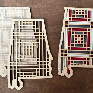 May include: Wooden coasters in the shape of the state of Alabama. One coaster is a natural wood color, while another features a geometric design with red, white, and gray accents. The coasters are on a brown surface.