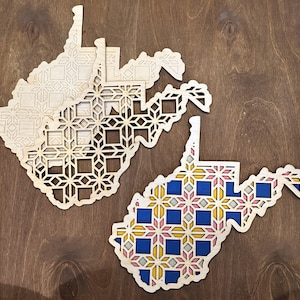 May include: Wooden laser-cut map of West Virginia with intricate geometric patterns. One map is natural wood, while another features blue, yellow, and pink accents. The maps are displayed on a dark wood surface.