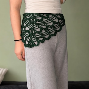 Könnte beinhalten: Ein dunkelwaldgrünes Häkel-Hüfttuch mit einem komplizierten, durchbrochenen Spitzenmuster und gewellten Kanten. Das dreieckige Accessoire ist aus Garn gefertigt und zeigt ein detailliertes, netzartiges Design, das über der Taille liegt.