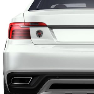 May include: A black and red car emblem with a shield design. The emblem is attached to the back of a white car.