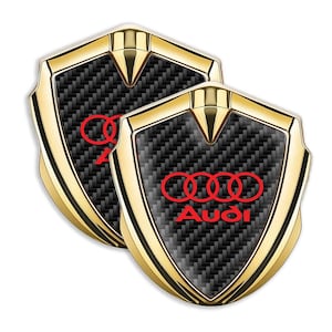May include: Two gold-colored car emblems with a carbon fiber pattern and a red Audi logo. The logo features four interlocking rings and the word "Audi" in red.