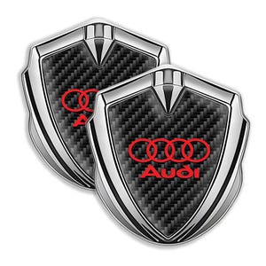 May include: Two silver and black carbon fiber car emblems with the Audi logo in red.
