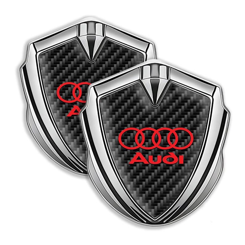 May include: Two silver and black carbon fiber car emblems with the Audi logo in red.