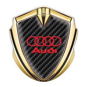 May include: A gold-colored car emblem with a black carbon fiber background. The emblem features four interlocking rings in red and the word "Audi" in red.