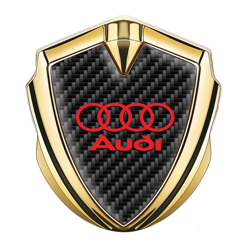 May include: A gold-colored car emblem with a black carbon fiber background. The emblem features four interlocking rings in red and the word "Audi" in red.